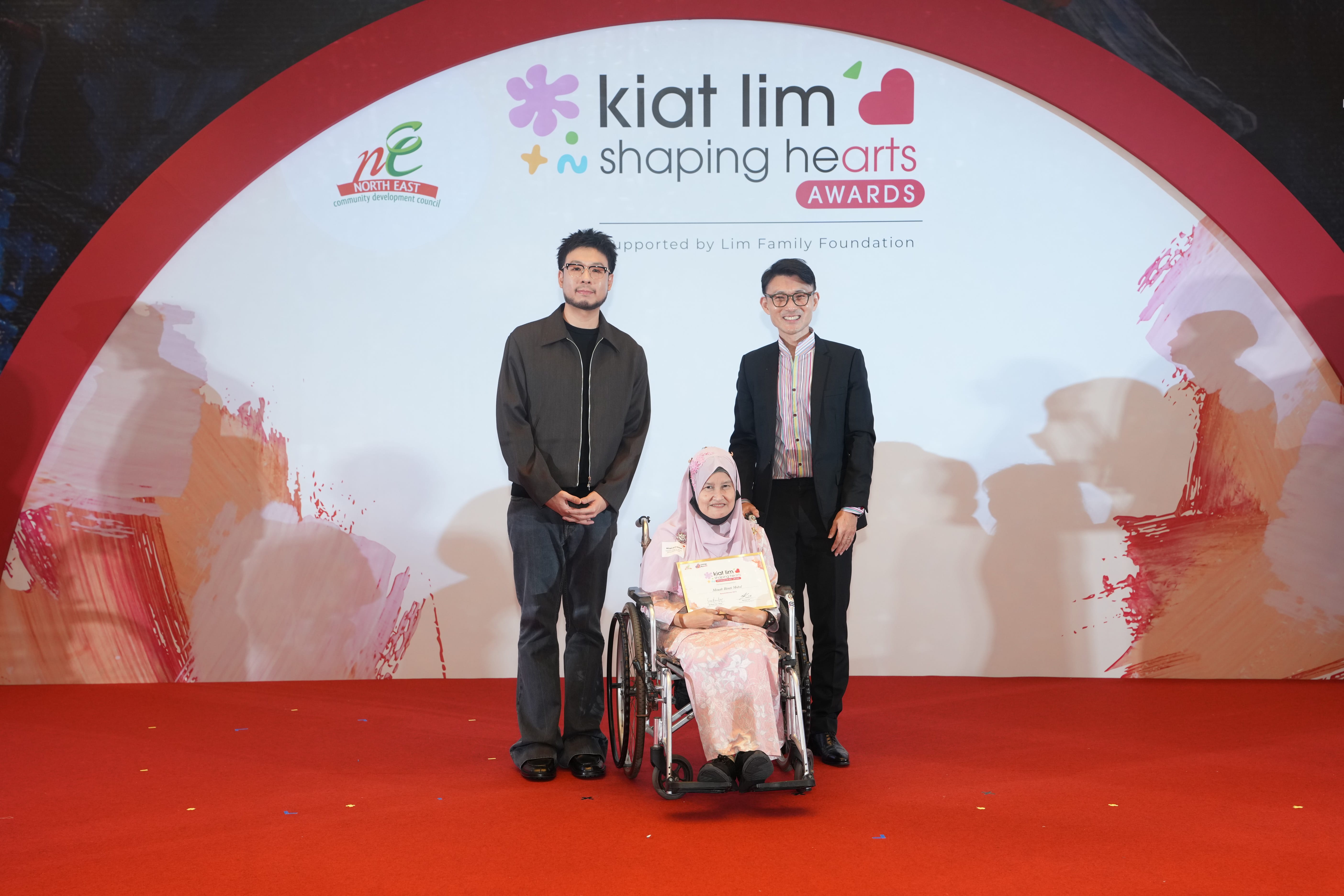 Award recipient standing on stage with the presenter, holding their certificate and posing for a group photo during the award ceremony, with a backdrop reading ‘Kiat Lim-Shaping Hearts Awards'.